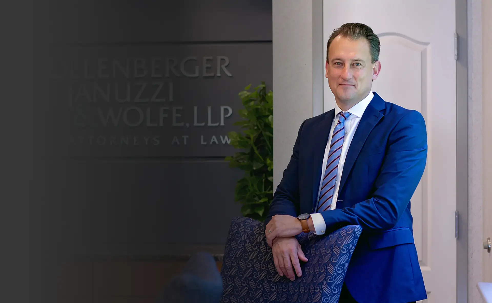 A professional man in a blue suit leans against a patterned chair, with a law firm sign visible in the background.