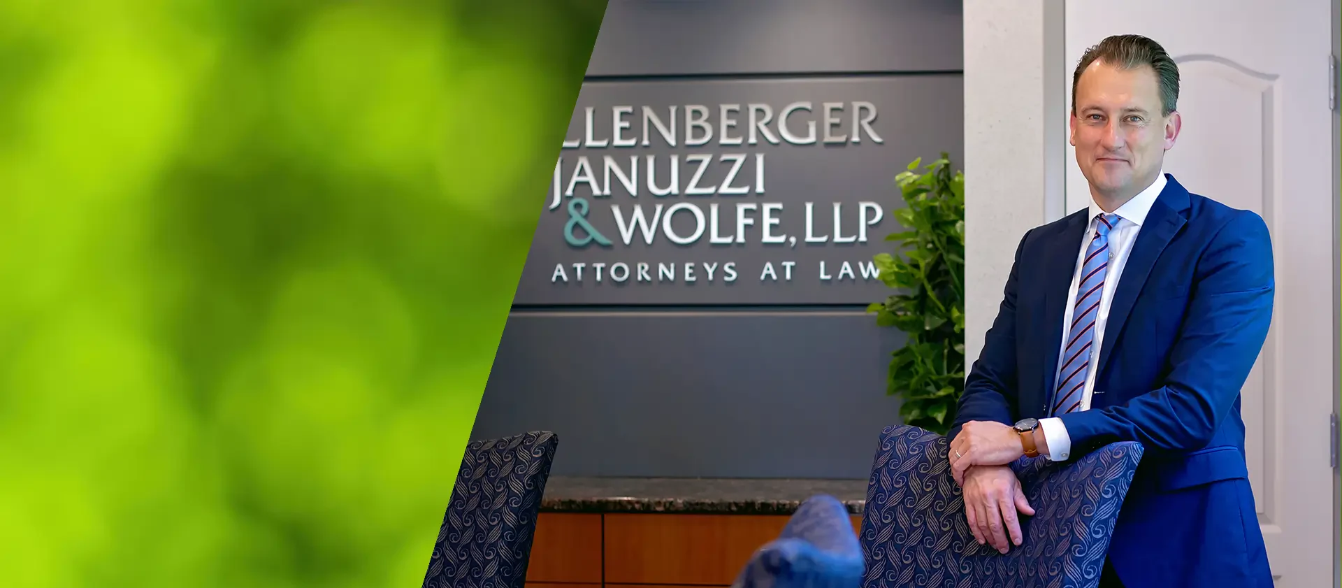 A professional attorney stands next to a dark sign reading 'Shollenberger Januzzi & Wolfe, LLP,' in a modern office environment