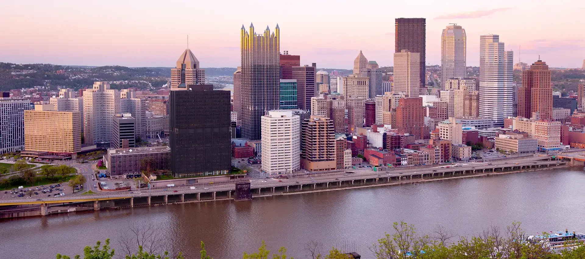 A panoramic view of Pittsburgh's skyline at dusk, showcasing tall buildings along the river, with soft pastel skies in the background